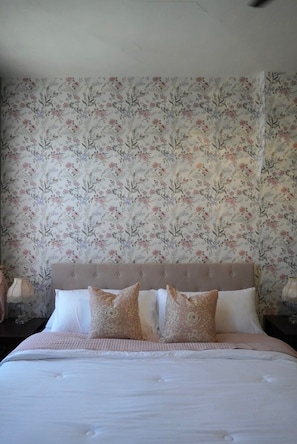 A bedroom with a floral wallpaper, a tufted headboard, and a bed with white bedding and patterned pillows.