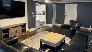 A living room with a flat-screen TV, leather sofas, a coffee table, a ceiling fan, and a kitchen in the background.