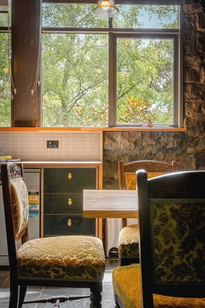A dining area with a view of trees through large windows, a stone wall, and a wooden table with chairs.