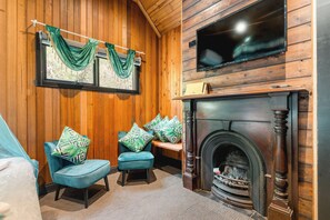 A room with wooden walls, a fireplace, two blue armchairs with patterned cushions, and a television mounted above the fireplace.