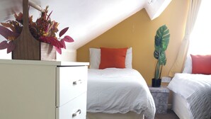 Ett sovrum med en vit byrå, två sängar med orange kuddar, en växt och en taklampa.