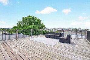 Eine Holzterrasse mit einer grauen Couch und einem weißen Teppich, mit Blick auf eine Grünanlage mit einem Baum und entfernte Gebäude.