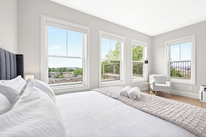 A modern hotel room with a large bed, multiple windows, a chair, and a view of the city. - East Boston Penthouse with Views 2 BR, 2BA (Boston)