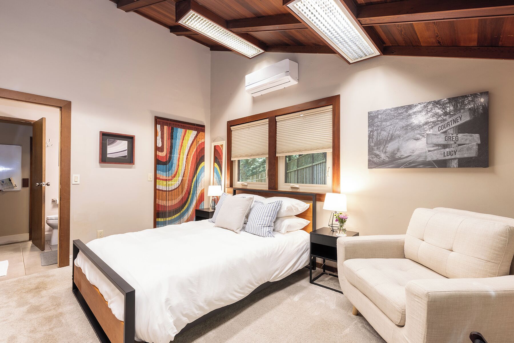 A bedroom with a bed, bedside table, chair, artwork, and a window with blinds.