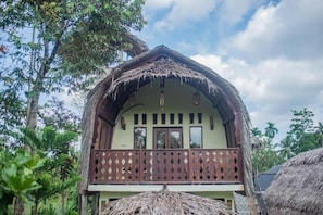 A traditional thatched-roof house with a balcony, surrounded by lush greenery.