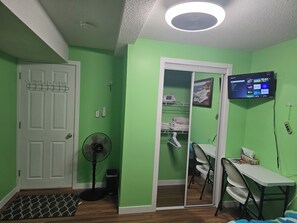Ein Zimmer mit grüner Wand, einer weißen Tür, einem Deckenventilator, einem an der Wand montierten Fernseher und einem Schreibtisch mit Stühlen.