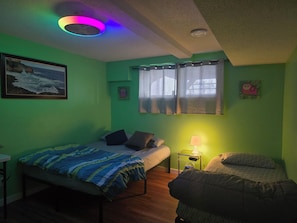 Una habitación con dos camas, ventilador de techo, ventana con cortinas, lámpara en una mesita de noche y un cuadro en la pared.