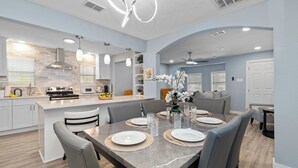 Una cocina moderna con una isla central, zona de comedor con una mesa de mármol y sala de estar con sofá y plantas decorativas.