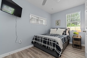 Een slaapkamer met een bed, een aan de muur bevestigde televisie, een raam met zonwering, een nachtkastje met een lamp en een deur.
