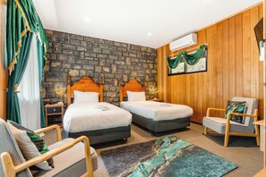 Hotellihuone, jossa on kaksi sänkyä, puinen seinä, kivestä tehty koristeellinen seinä, vihreä verho, tuoli ja pieni pöytä.