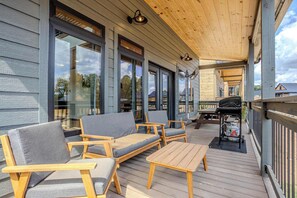 Eine überdachte Terrasse im Freien mit Holzmöbeln, einem Grill und großen Fenstern.