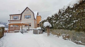 Una casa de dos pisos con techo nevado, cerca de madera y un muro de piedra en un lateral.