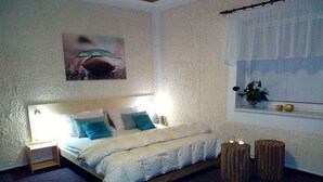 Un dormitorio con cama, mesitas de noche, una ventana con persianas, una planta y un cuadro en la pared.
