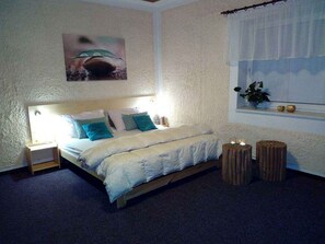 Un dormitorio con cama, mesitas de noche, una ventana con persianas, una planta y un cuadro en la pared.