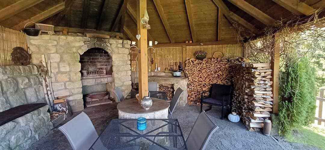 A rustic outdoor seating area with a stone fireplace, wooden furniture, and a thatched roof.