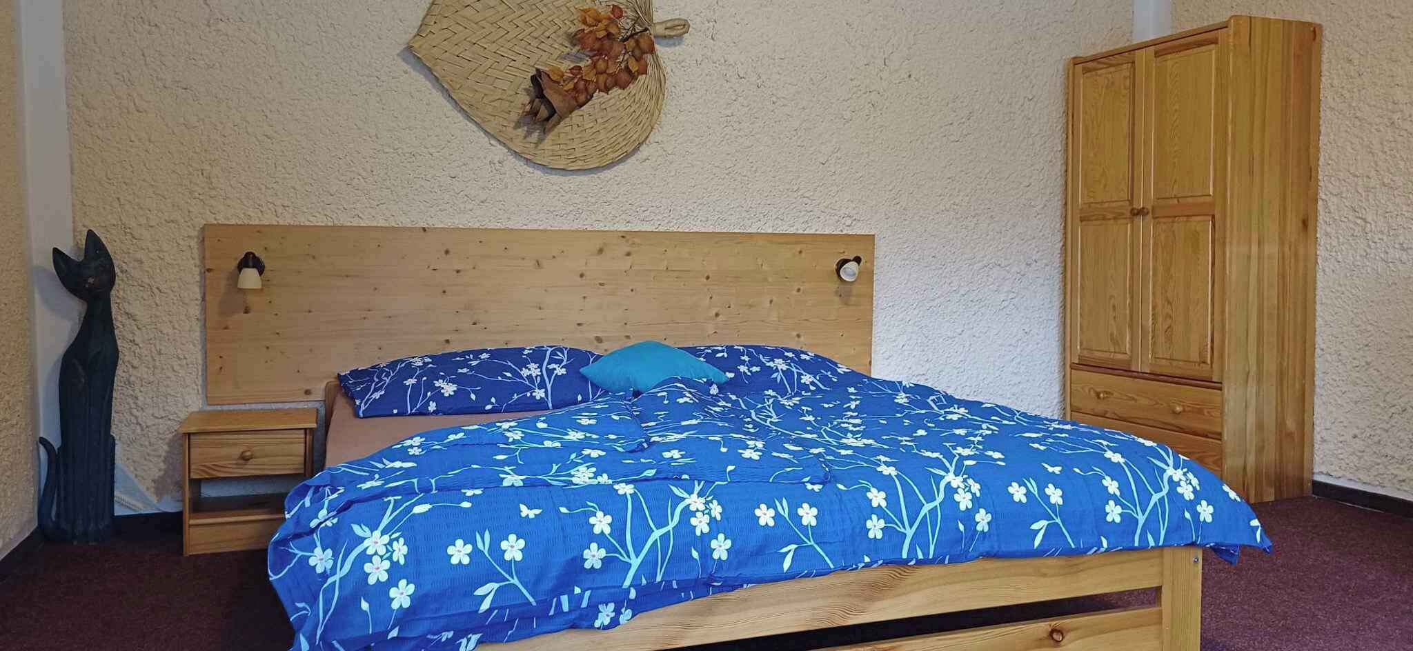 A wooden bed with a blue floral bedspread and a matching pillow, a wooden nightstand, a small wall decoration, and a wooden wardrobe.