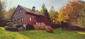 Ein Holzhaus mit geneigtem Dach, umgeben von einem gepflegten Rasen und Bäumen in Herbstfarben.