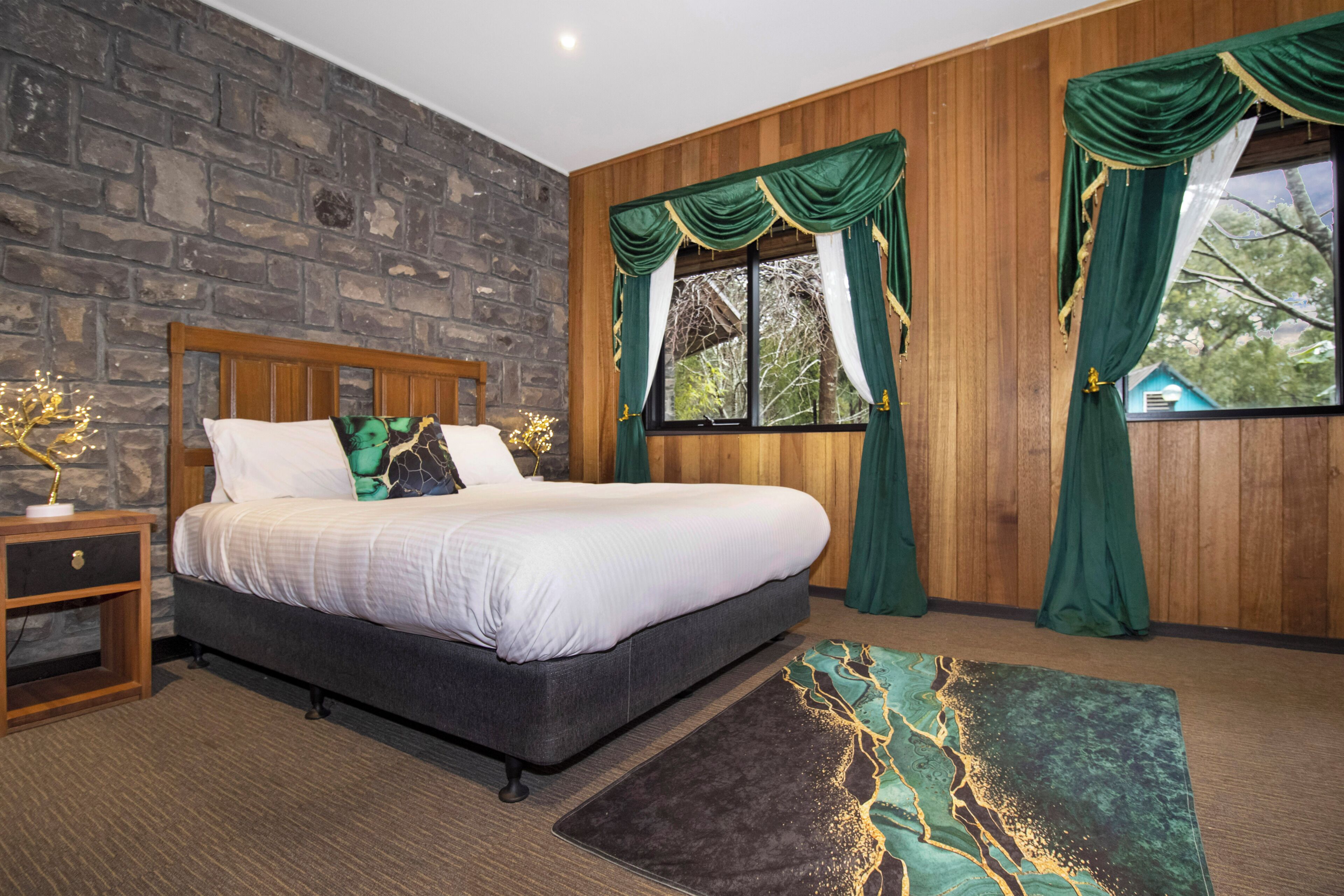 Une chambre avec un mur en pierre, un lit doté d’une tête de lit en bois, des rideaux verts et une vue sur les arbres par la fenêtre.