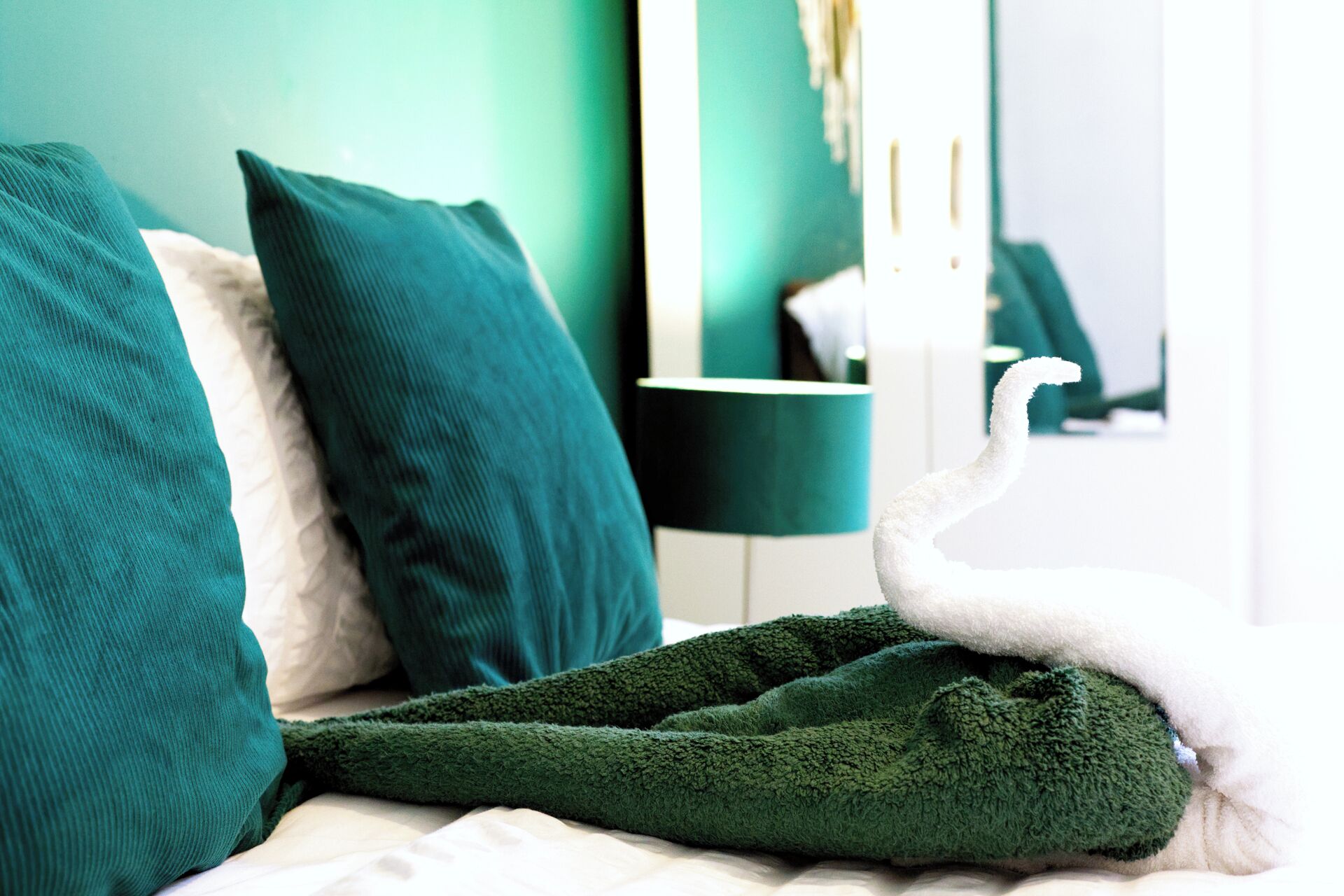 Una cama con una manta y almohadas verdes, una mesita de noche con una lámpara, y un espejo que refleja la habitación.