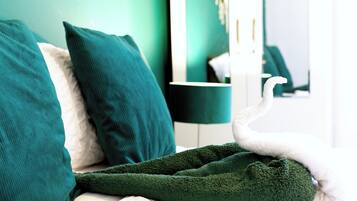 Una cama con una manta y almohadas verdes, una mesita de noche con una lámpara, y un espejo que refleja la habitación.