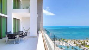 Een balkon met een eettafel en stoelen, met uitzicht op een strand met palmbomen en een zwembad.