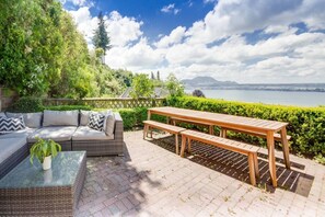 An outdoor patio with a sofa, a wooden table, and benches, overlooking a lake and mountains. - Serendipity Too- panoramic lake views, and contemporary interiors (Taupō)