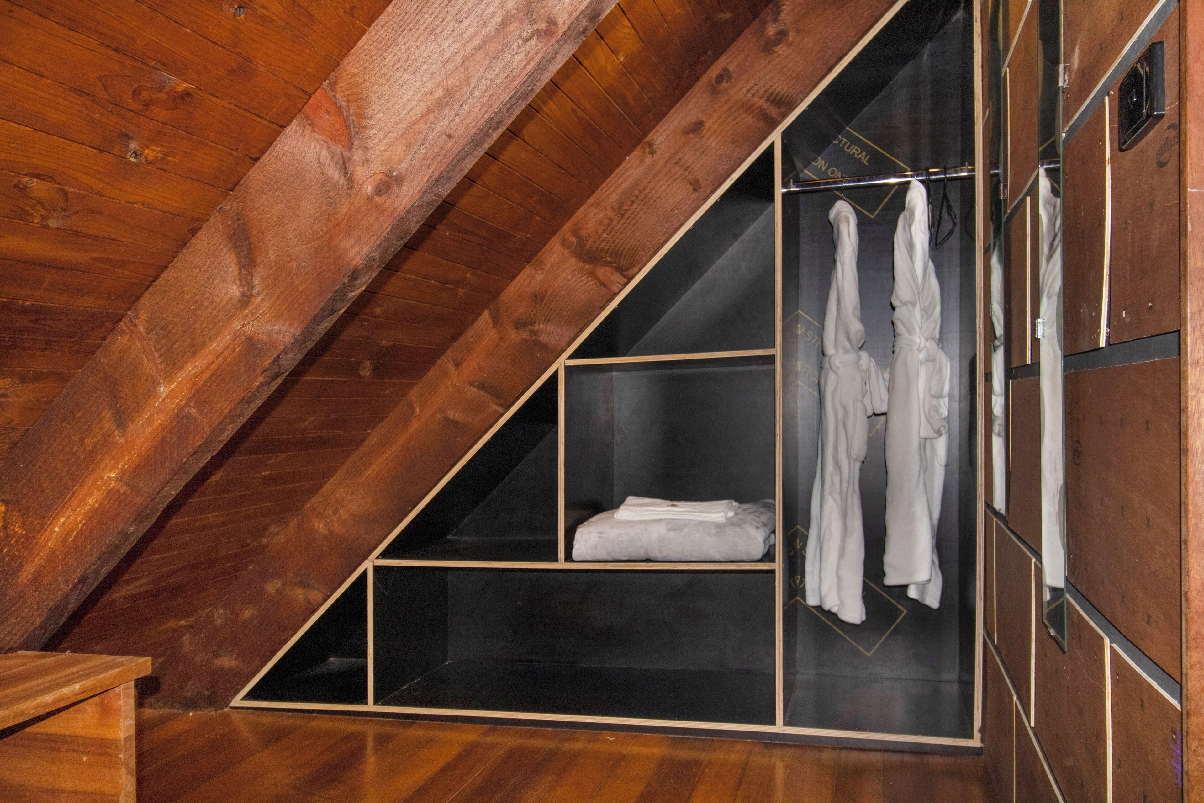 Une chambre mansardée triangulaire avec des poutres en bois, un placard intégré avec une tringle pour suspendre les vêtements et une étagère avec des serviettes pliées.