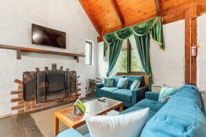 A living room with a fireplace, a sofa set, a television, and a window with curtains. - Myrtle Loft - Sun-Kissed Forest Garden, Emerald (Emerald)