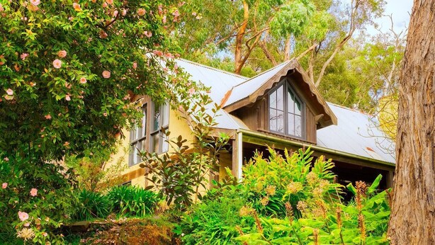 Une maison de campagne au toit à pignon, entourée d’une végétation luxuriante et de plantes en fleurs.