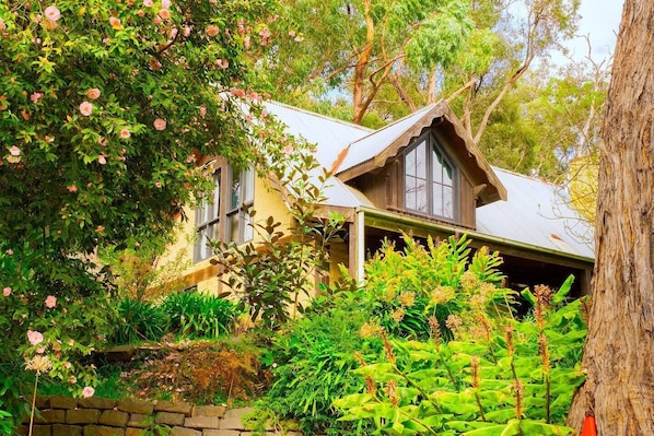 Une maison de campagne au toit à pignon, entourée d’une végétation luxuriante et de plantes en fleurs.