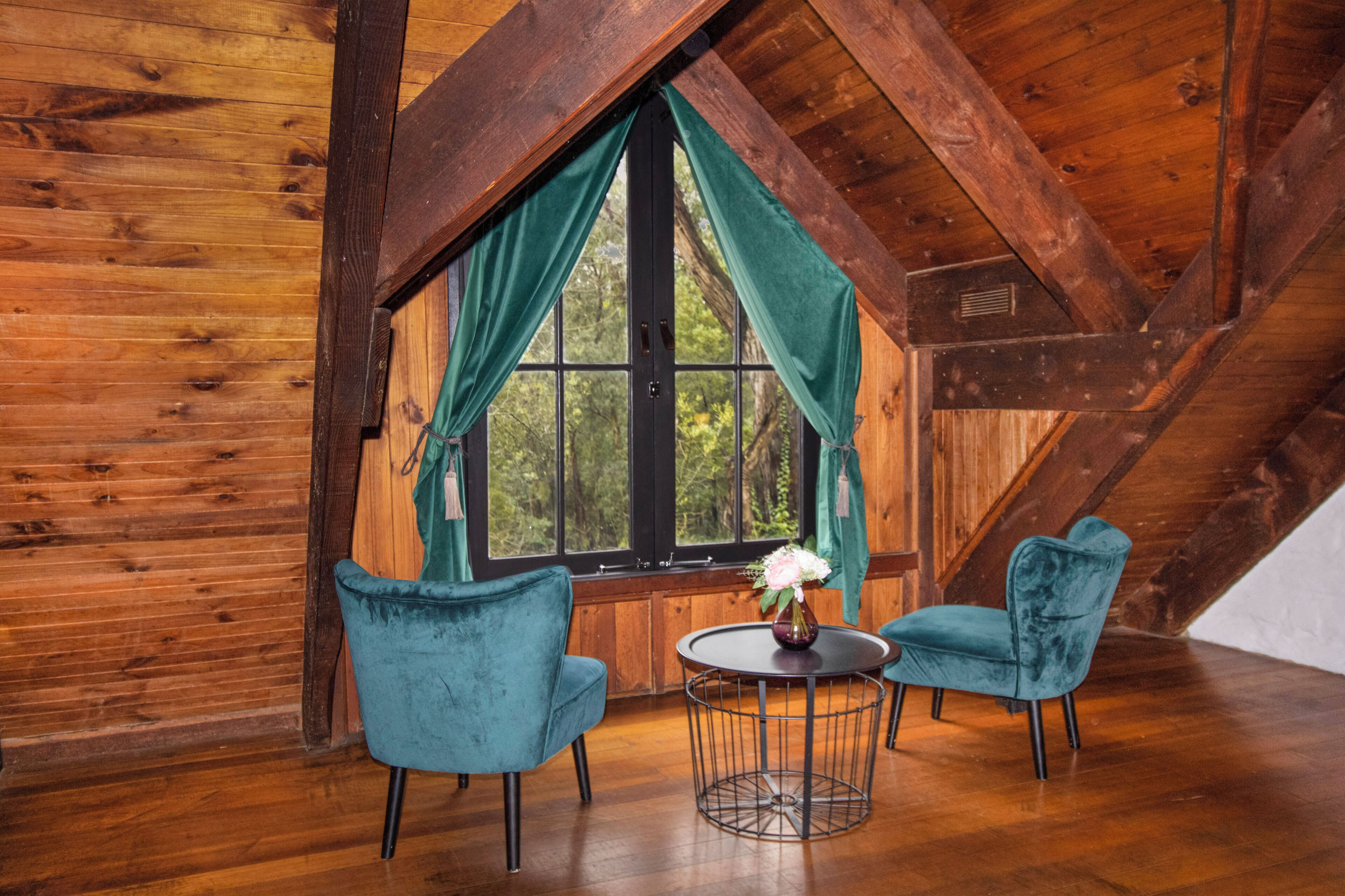 A room with wooden walls, a large window with curtains, two chairs, a small table, and a vase with flowers.