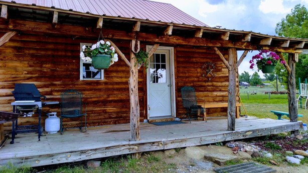 A rustic log cabin with a porch, a grill, and hanging flower baskets.