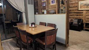 A rustic dining area with a wooden table and chairs, a small kitchen area with a refrigerator, and a wall with framed pictures and labels.