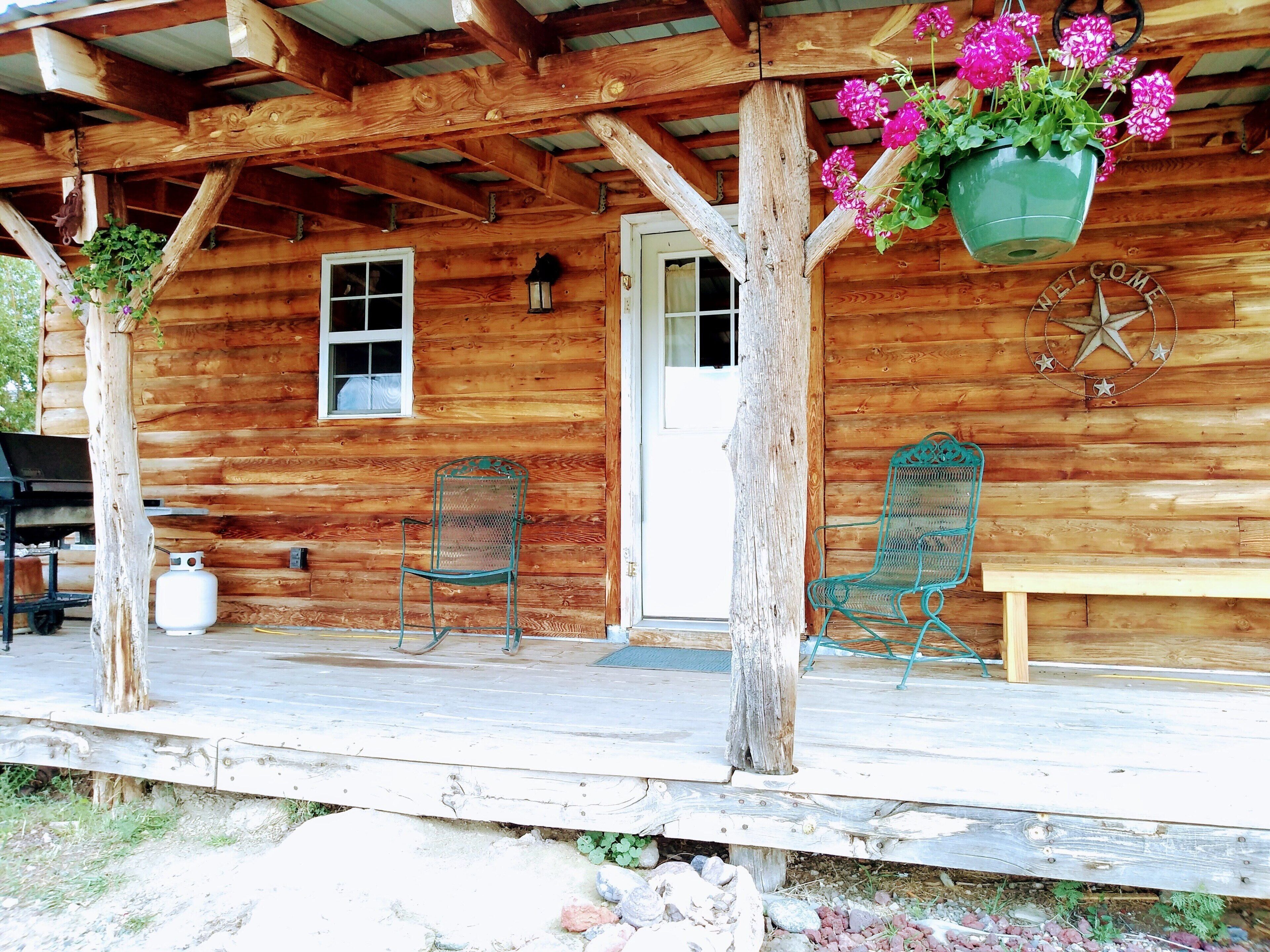A wooden cabin with a porch, two metal chairs, a bench, and a hanging flower pot.