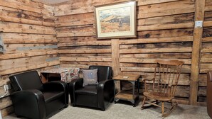 A room with wooden walls, a framed picture, a small table with a cushion, two chairs, and a rocking chair.