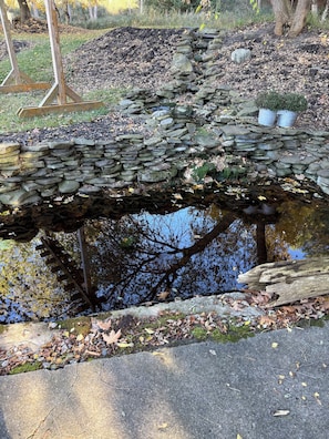 A stone bridge over a pond with a wooden structure on one side, surrounded by trees and a grassy area.
