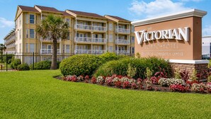 A multi-story building with balconies, a sign for 'The Victorian Condominium Hotel,' and a well-maintained lawn with flower beds. - Coral Reef Retreat | Beachfront Escape (Galveston)
