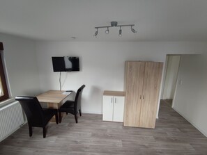 Ein Zimmer mit Esstisch, Stühlen, einem an der Wand befestigten Fernseher, einem Kleiderschrank und einer Deckenleuchte.