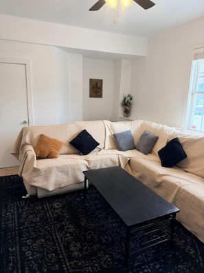Een woonkamer met een bank, een salontafel, een gekleurd vloerkleed, een plafondventilator en een raam met jaloezieën.