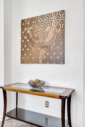 Ein Wandbild mit geometrischem Muster, ein Konsolentisch mit Glasplatte und einer dekorativen Schale sowie eine Steckdose an der Wand.