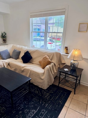 Ein Wohnzimmer mit einer Couch, einem kleinen Tisch, einer Lampe und Blick ins Freie.