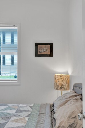 Ein Schlafzimmer mit einem Bett, einem Fenster mit Jalousien, einer Wandlampe und einem gerahmten Bild.