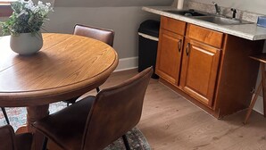 Ein Essbereich mit einem runden Holztisch, zwei Stühlen, einem Blumentopf sowie eine Küche mit Holzschränken und einem Spülbecken.