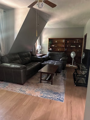 A living room with a sectional sofa, a coffee table, a wooden floor, a ceiling fan, and a bookshelf.