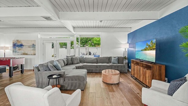 Un salon moderne avec un canapé d’angle, un pouf et un téléviseur posé sur un meuble en bois.