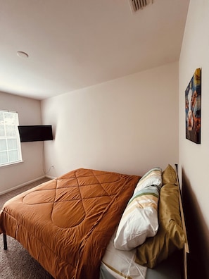 Ein Schlafzimmer mit Bett, einem an der Wand befestigten Fernseher und einem Fenster mit Jalousien.