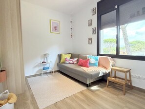 Ein modernes Wohnzimmer mit einem grauen Sofa, farbenfrohen Kissen, einem kleinen Holzschemel, einem weißen Beistelltisch mit Lampe und einem großen Fenster mit Blick auf Bäume.