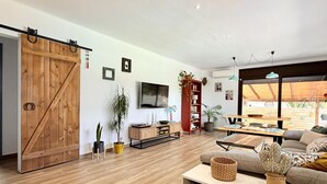 Ein geräumiger Wohnbereich mit einer Schiebetür im Scheunenstil, Holzböden, einer Couch, einem Fernseher und einem Essbereich mit Tisch und Stühlen.