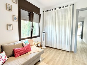 Un salón con sofá, un radiador portátil, una ventana con persianas y una puerta que da a otra habitación.