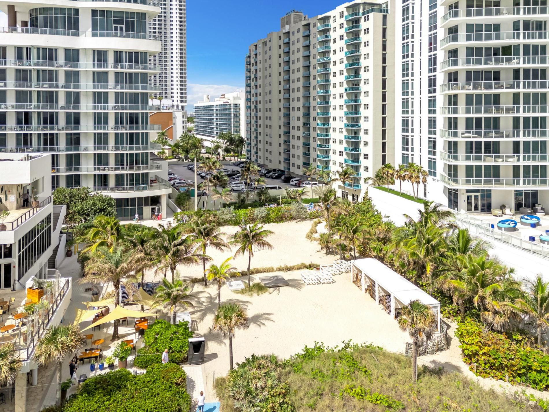 A high-rise residential building with multiple balconies, a sandy beach area with palm trees, and a parking lot.